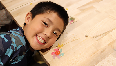 a child smiles at the camera next to their jewelry project banner