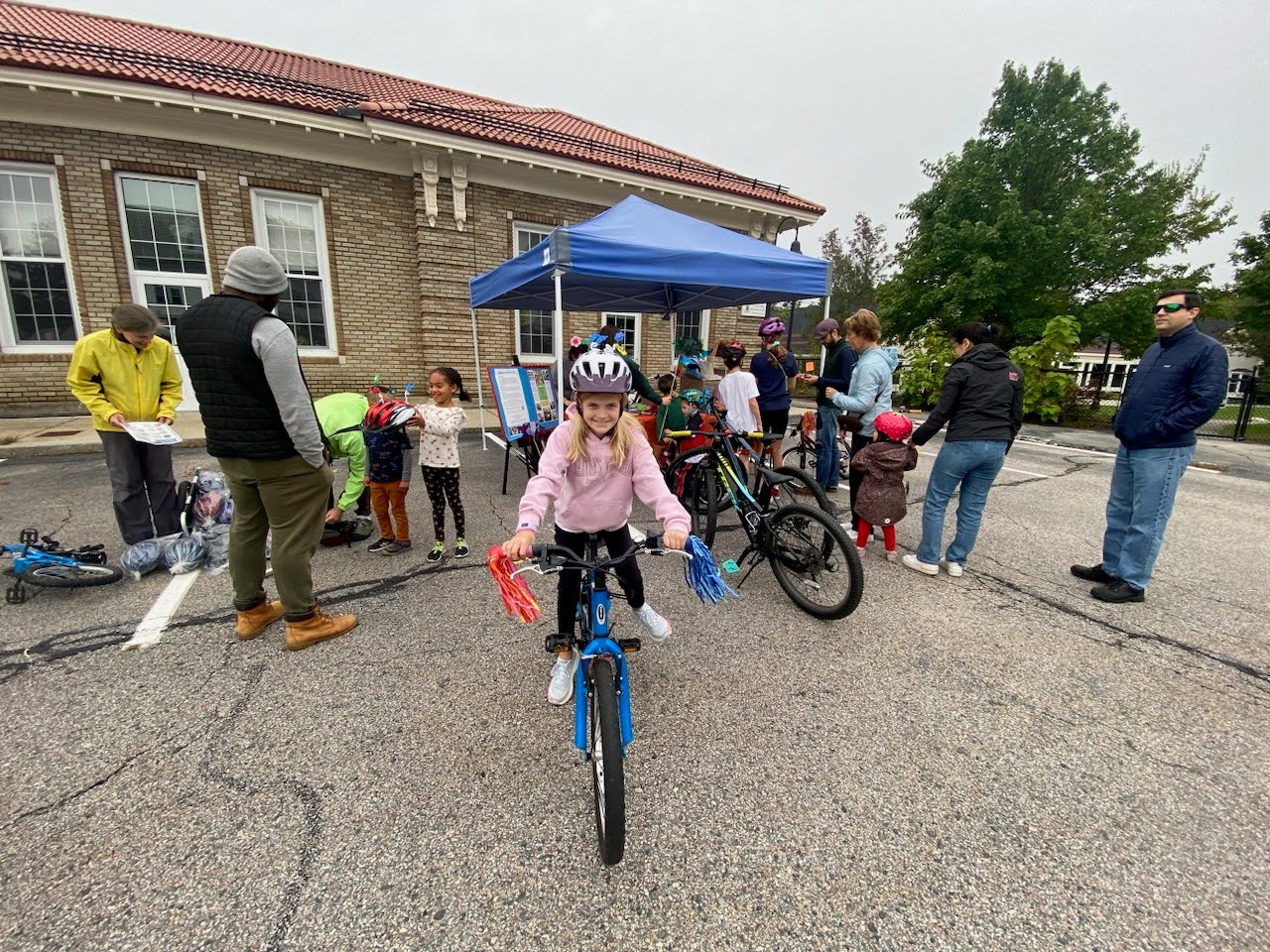 a smiling girl on a bicycle next to a pop up tent with other bicyclists by the Fowler Branch Library